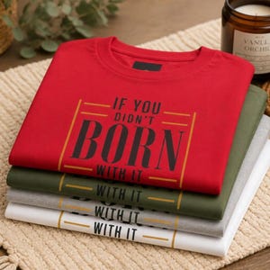 Può includere: Una pila di quattro magliette a girocollo piegate in rosso, verde oliva, grigio melange e bianco. La maglietta rossa in alto presenta il testo nero "IF YOU DIDN'T BORN WITH IT" incorniciato da due linee dorate, mentre quelle sotto riportano "WITH IT".