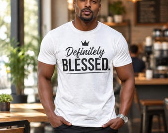 May include: A white short-sleeved t-shirt featuring a black graphic print on the chest. The design includes a small crown icon above the text "Definitely BLESSED." written in a combination of script and bold sans-serif fonts.