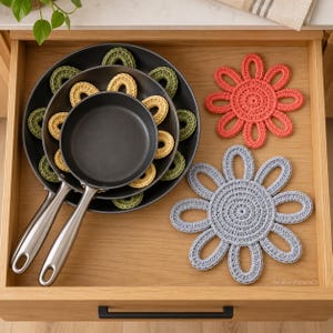 Op de afbeelding: Een houten lade met drie in elkaar gestapelde zwarte koekenpannen met zilveren handgrepen, gescheiden door kleine olijfgroene en gele gehaakte ringen. Rechts liggen twee grote, bloemvormige gehaakte onderzetters in koraal en lichtblauw.