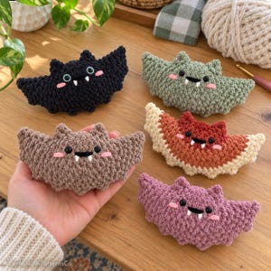 May include: Five crocheted bat plushies in black, sage green, light brown, ombre orange and cream, and dusty pink. Each bat features black safety eyes, two small white fangs, and pink embroidered cheeks. The texture is consistent across all items.