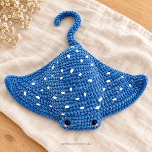 May include: A blue crochet stingray plush toy featuring a textured yarn surface with white embroidered spots, two black plastic eyes, and small pink stitched cheeks. A curved tail extends from the top of the body. Text reads YarnBloomPatternsCo.
