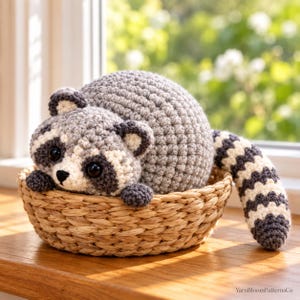 May include: A round, crocheted raccoon plush toy with gray and white yarn, featuring black eye patches, a small black nose, and a striped tail, resting inside a woven tan basket. The text YarnBloomPatternsCo is visible in the bottom right corner.