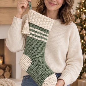 May include: A crochet Christmas stocking featuring a sage green body with cream-colored accents on the toe, heel, and ribbed cuff. The design includes two horizontal cream stripes below the cuff and a decorative cream tassel attached to the top.