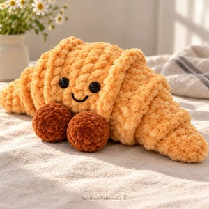 May include: A plush croissant toy made of light orange yarn with a textured, looped finish. It features two black plastic safety eyes, a stitched black smile, and two small, round, dark brown yarn feet. The text YarnBloomPatternsCo is visible.