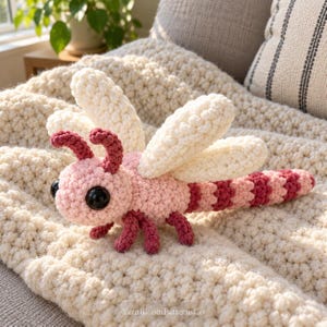 May include: A crocheted dragonfly toy featuring a light pink head and body, dark pink antennae and legs, and four large, textured off-white wings. The body has alternating dark pink stripes. It has large black eyes and the text YarnBloomPatternsCo.
