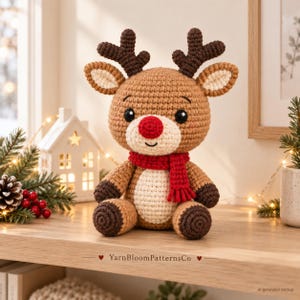 Peut inclure: Une peluche renne au crochet avec un corps marron clair, des bois marron foncé et un ventre crème. Le renne présente un nez rouge vif, des yeux noirs et une écharpe rouge autour du cou. Le texte YarnBloomPatternsCo est visible.