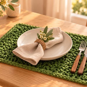 Può includere: Una tovaglietta rettangolare all'uncinetto di colore verde con un motivo a maglia spessa e strutturata. Un piatto in ceramica bianca poggia sulla tovaglietta, contenente un tovagliolo di lino beige con un anello.