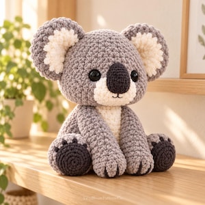 May include: A crocheted koala stuffed animal featuring a gray body, large round ears with white centers, a white chest patch, and a dark gray nose. The koala has black safety eyes and small dark gray feet with visible toe details. The text YarnBloomPatternsCo is visible at the bottom.
