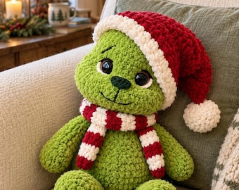 May include: A green crocheted plush toy featuring a dark green nose, large black eyes, and a stitched mouth. The toy wears a red and white crocheted Santa hat with a pom-pom and a matching red and white striped scarf. The text YarnBloomPatternsCo is visible.