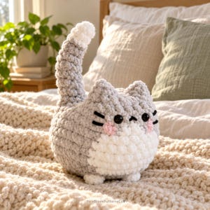 May include: A plush crochet cat toy featuring a round grey body, a white belly, and a long upright tail with a white tip. The cat has black plastic eyes, black embroidered whiskers, and small pink stitched cheeks. The text YarnBloomPatternsCo is visible.