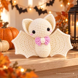 May include: A cream-colored crocheted bat plush toy featuring large pointed ears, black safety eyes, pink stitched cheeks, and a small pink bow tie at the neck. The bat has wide, scalloped wings and a rounded body, crafted with visible yarn stitches.