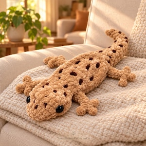 Peut inclure: Un jouet en peluche lézard crocheté de couleur beige avec des taches marron foncé et des yeux en plastique noir. Le lézard a un aspect texturé et doux avec quatre pattes et une longue queue. Le texte YarnBloomPatternsCo est visible en bas.