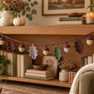 May include: A crochet garland featuring alternating oak leaves and acorns in shades of sage green, cream, light purple, and dark brown, strung along a dark brown yarn cord. The decorative piece displays various textured leaf shapes.