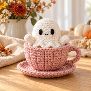 May include: A white crocheted ghost figure with black eyes and pink cheeks sitting inside a dusty pink crocheted teacup and saucer set. The ghost has a rounded head and small arms, while the teacup features a ribbed texture and a curved handle.