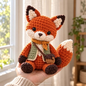 May include: A small, handcrafted crochet fox plush toy featuring orange, white, and dark brown yarn. The fox wears a sage green vest, a mustard yellow scarf, and a small brown acorn-shaped bag. The text YarnBloomPatternsCo is visible.