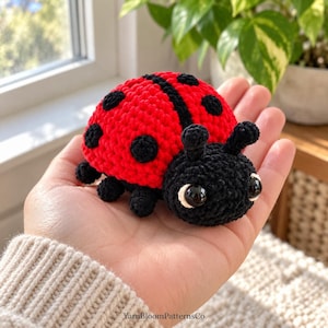 May include: A small, handmade crochet ladybug toy featuring a bright red back with black spots and a solid black head. The plush insect has two black antennae, six small black legs, and two large, glossy black eyes with white borders. The text YarnBloomPatternsCo is visible at the bottom.