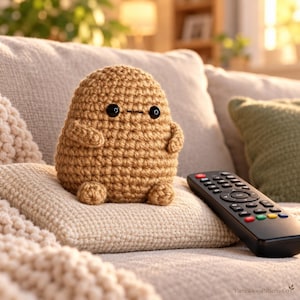May include: A tan crochet plush toy shaped like a rounded potato with two small black plastic eyes and a simple stitched mouth. The figure has two small arms and two small feet, sitting on a textured beige cushion next to a black television remote.