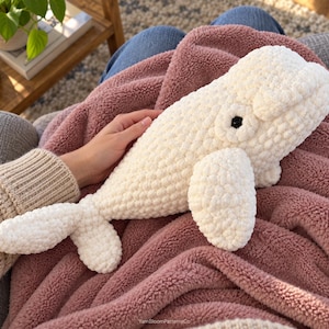 May include: A white crocheted beluga whale plush toy made from soft, chunky yarn. The whale features a rounded head, a prominent forehead, small side fins, a tail fluke, and a single black plastic eye. The text YarnBloomPatternsCo is visible.