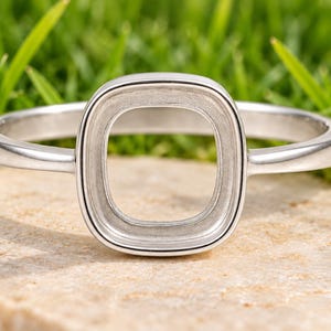 Könnte beinhalten: Ein silberfarbener Metallring mit einer mittigen, leeren, abgerundet quadratischen Fassung. Die Ringschiene ist glatt und poliert und führt zu einem vertieften, mattierten Innenbereich innerhalb des quadratischen Rahmens für einen Stein.