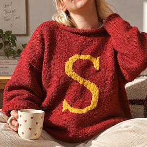 Puede incluir: Un suéter de punto de color rojo intenso con mangas largas y una letra S amarilla texturizada grande centrada en la parte delantera. El suéter tiene un cuello ancho y redondeado con una textura de punto grueso. Una taza de cerámica blanca con corazones rojos.