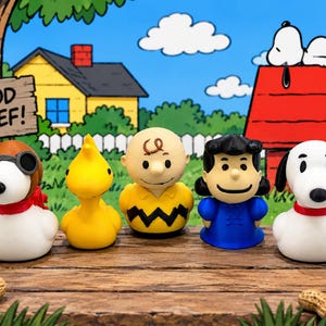 May include: A set of five Peanuts character figurines including a Snoopy aviator, a yellow bird, Charlie Brown, Lucy, and a standard Snoopy. The figures are arranged on a wooden surface in front of a cartoon backdrop with the text GOOD GRIEF.