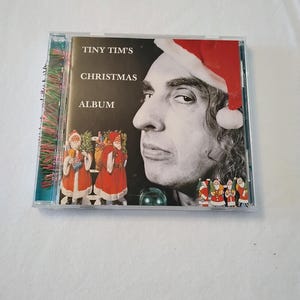 Può includere: Un CD di Tiny Tim's Christmas Album in una custodia di plastica trasparente. La copertina presenta un ritratto in bianco e nero di Tiny Tim con un cappello da Babbo Natale rosso, il testo "TINY TIM'S CHRISTMAS ALBUM" e illustrazioni di Babbo Natale.