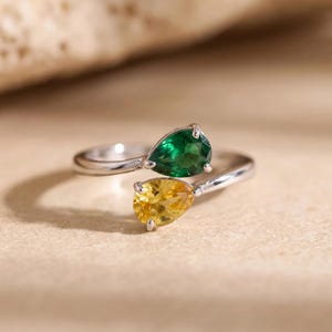 Puede incluir: Un anillo abierto de metal plateado pulido, con dos piedras preciosas en forma de pera. Una piedra es de color verde esmeralda intenso, mientras que la otra es de un amarillo dorado brillante. Las piedras están sujetas por garras.