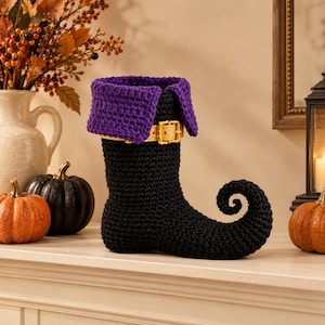 May include: A black crocheted boot with a curled toe, featuring a wide purple crocheted cuff at the top and a tan strap with a gold-colored buckle wrapped around the ankle area of the boot. The item is constructed from yarn.