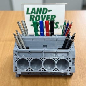 Puede incluir: Un organizador de escritorio gris impreso en 3D con forma de bloque de motor con múltiples ranuras cilíndricas que contienen varios bolígrafos y rotuladores. Detrás hay un letrero blanco con el texto verde LAND ROVER PARTS.IT.