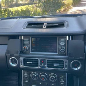 May include: A black vehicle dashboard featuring a central infotainment screen, air conditioning vents, and a control panel with multiple circular dials and buttons. The console includes a start-stop button, navigation controls, and phone settings.