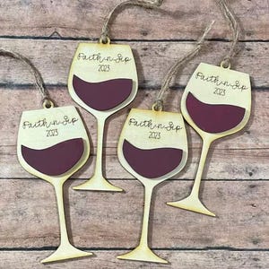 May include: Four wine glass ornaments made of light-colored wood with a dark maroon inlay representing wine. Each ornament features the text Faith n Sip 2023 in black script and includes a rustic twine loop for hanging on a tree or display.