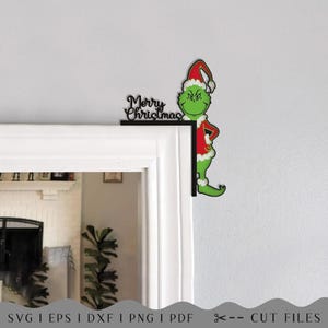 May include: A black corner door frame decoration featuring a green character wearing a red and white Santa hat and suit, with the text Merry Christmas in a cursive font. The design is intended for placement on the top corner of a door frame.