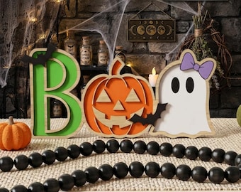 May include: A wooden Halloween sign spelling BOO with a green letter B, an orange jack-o'-lantern pumpkin, and a white ghost with a purple bow. The sign features black bat silhouettes and is displayed with a black wooden bead garland.