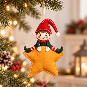 Pode incluir: Um enfeite de árvore de Natal em feltro com um pequeno elfo de gorro vermelho, camisa verde e mangas listradas, sentado sobre uma grande estrela amarela de cinco pontas. O ornamento é costurado com linha branca.