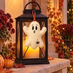 May include: A black metal lantern with glass panels containing a glowing pillar candle. A cream-colored felt ghost ornament with a surprised expression and a small black spider graphic hangs from the top handle by an orange ribbon.