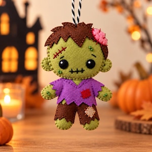 Può includere: Un ornamento in feltro a forma di zombie con viso verde chiaro, occhi neri, bocca cucita e capelli castani. La figura indossa una maglia viola con una toppa rossa e pantaloni marroni con una toppa beige, appesa a un cordino a righe bianche e nere.
