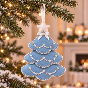 May include: A light blue felt Christmas tree ornament featuring three scalloped tiers. The tree is decorated with white stitched chain patterns, small white faux pearls, and a white ribbon bow at the top, finished with a white felt star.