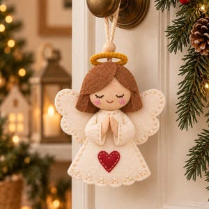 May include: A felt angel ornament featuring a cream-colored dress with a red heart appliqué, brown hair, a yellow halo, and white wings. The figure has closed eyes, pink cheeks, and is suspended by a braided cord from a brass-colored door handle.