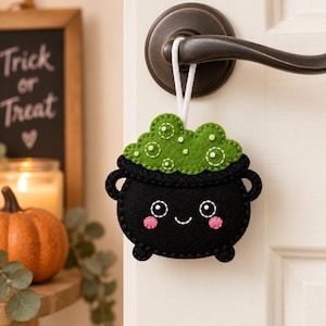 May include: A black felt cauldron ornament with a smiling face, pink cheeks, and green bubbling liquid at the top. The ornament features white stitching details and a white cord loop for hanging, suspended from a dark metal door handle.