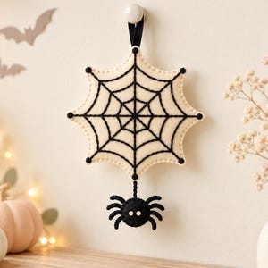 May include: A cream-colored felt spiderweb ornament with black stitched web details, hanging from a black ribbon. A small, round black felt spider with two white bead eyes is attached to the bottom of the web by a short black cord.