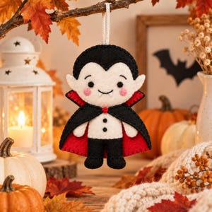 May include: A felt vampire ornament featuring a cream-colored face with pink cheeks, two small white fangs, and black hair. The figure wears a black cape with a bright red lining, a white shirt with two black buttons, and black trousers, suspended by a white cord.