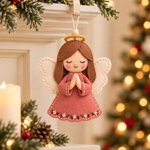 May include: A felt angel ornament featuring brown hair, a yellow halo, and a dusty rose dress with white and green floral embroidery. The angel has cream-coloured wings, closed eyes, and pink cheeks, with hands pressed together in a prayer position.