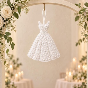 May include: A white hanging ornament shaped like a dress, featuring a sweetheart neckline and a flared skirt. The entire surface is embellished with small white beads and pearls, and it includes a white cord loop for hanging from a hook.