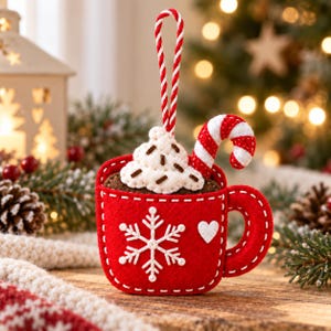 Könnte beinhalten: Ein roter Filz-Tassenanhänger mit einer weißen Schneeflocke und einem kleinen Herz auf der Vorderseite. Die Tasse ist mit weißer Filz-Schlagsahne, braunen Streuseln und einer rot-weiß gestreiften Zuckerstange gefüllt und hat eine Kordel.