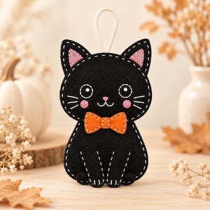 May include: A black felt cat ornament featuring white stitched outlines, pink embroidered cheeks, and a bright orange bow tie. The cat has large white eyes with black pupils, a small pink nose, and a white cord loop at the top for hanging.