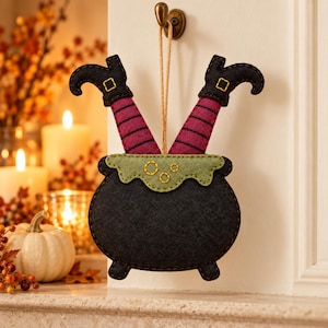 May include: A felt hanging decoration featuring a black cauldron with a light green rim and two legs extending upward. The legs are covered in magenta and black striped fabric, ending in black boots with yellow stitched buckles.