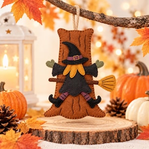 May include: A felt hanging ornament shaped like a brown tree trunk with a witch figure attached to the front. The witch wears a black hat, black dress, and purple striped leggings, holding a yellow broomstick. It features a tan twine loop.
