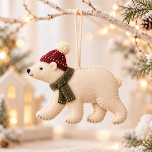 May include: A cream-colored felt polar bear ornament featuring a dark red knit-style hat with a small pom-pom and a dark olive green scarf. The bear is stitched with visible thread and includes a light-colored string loop for hanging.