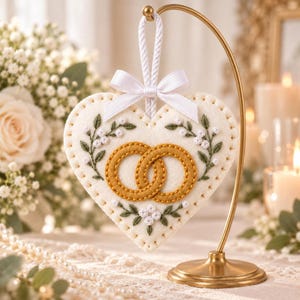 May include: A heart-shaped white felt ornament featuring two interlocking gold rings embroidered in the center, surrounded by green leaf and white flower embroidery. The ornament hangs from a white rope with a white satin bow on a gold stand.