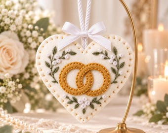 Op de afbeelding: Een hartvormig wit vilt ornament met twee in elkaar grijpende gouden ringen in het midden, omringd door groen blad- en wit bloemenborduurwerk. Het ornament hangt aan een wit koord met een witte satijnen strik aan een gouden standaard.